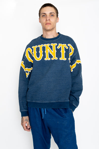 Kapital long sleeve sweatshirt pullover that is an indigo fleece loose fit. Loose fit. Across the front is a large graphic text of the KOUNTRY logo. The back has a T-shaped split back and a cinch back. Color: Indigo 100% Cotton. Made In Japan. Unisex. 