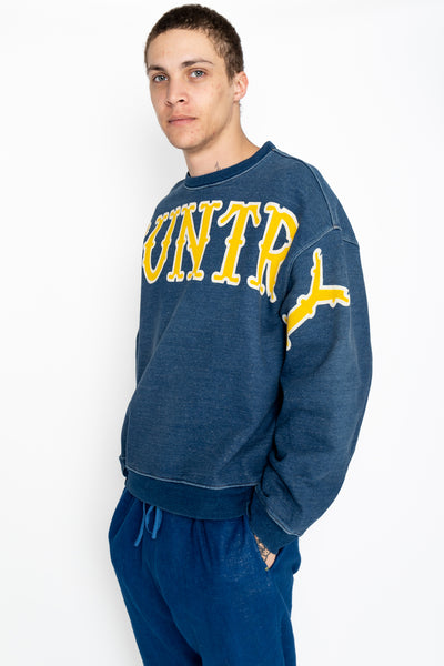 Kapital long sleeve sweatshirt pullover that is an indigo fleece loose fit. Loose fit. Across the front is a large graphic text of the KOUNTRY logo. The back has a T-shaped split back and a cinch back. Color: Indigo 100% Cotton. Made In Japan. Unisex. 